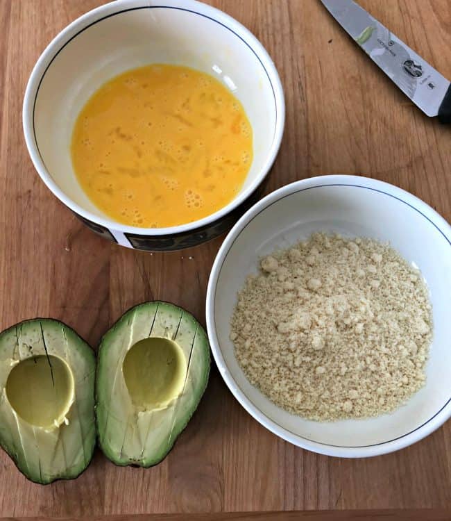 one bowl with beaten egg and another with almond meal next to a cut avocado