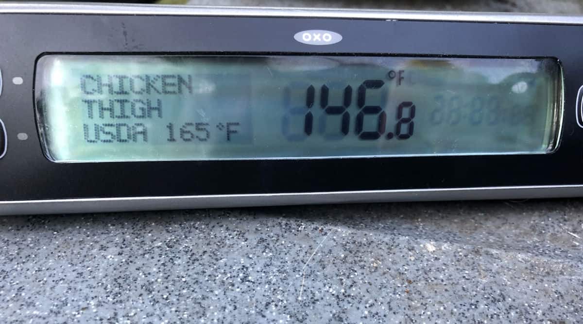 Digital meat thermometer showing the internal temperature of chicken legs cooking on the grill, nearing optimal doneness.