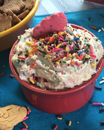 Dunkaroo Dip, also known as funfetti cake batter dip, in a small bowl garnished with sprinkles and surrounded by crackers and cookies for dipping