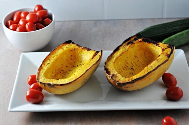 2 halves of grilled spaghetti squash on a white platter with grape tomatoes nearby