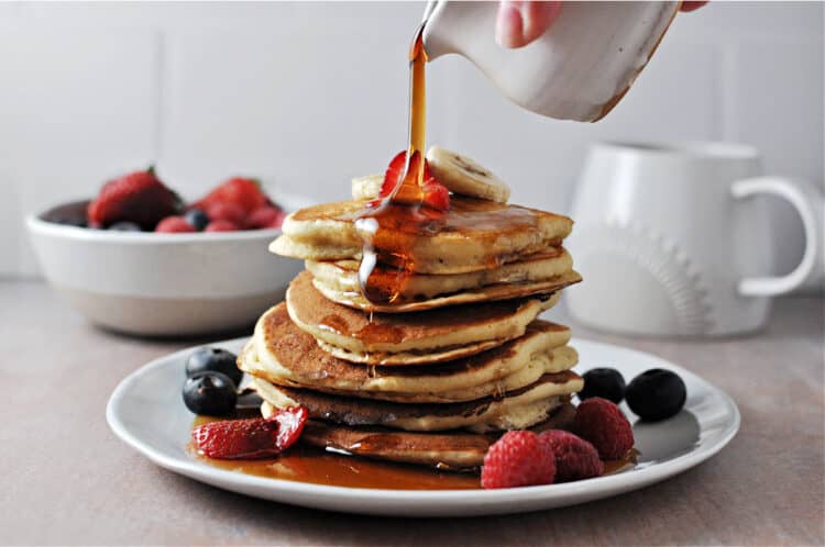 syrup being poured onto a stac of oat milk pancakes with fruit on the plate
