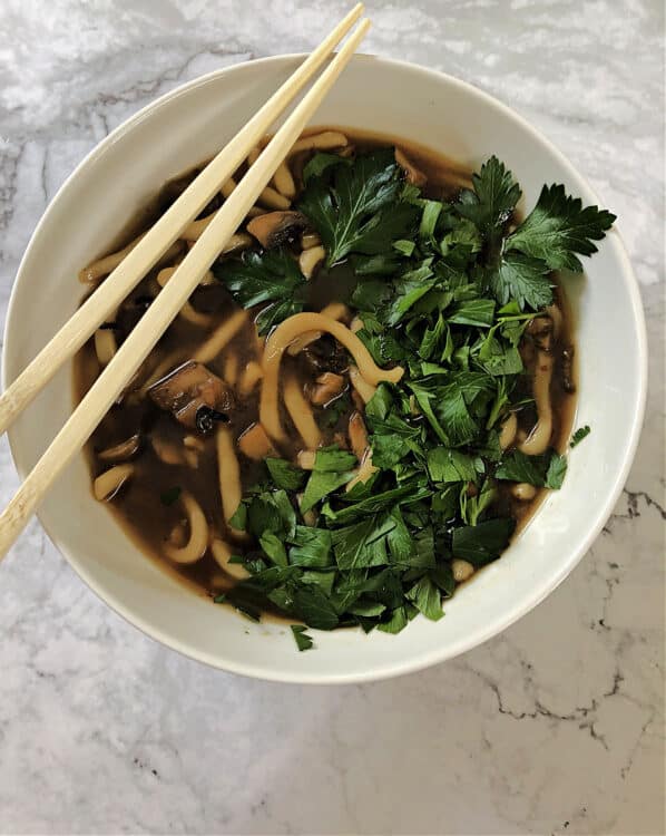 spicy mushroom noodle soup served in a white bowl with chopped frresh herbs on top