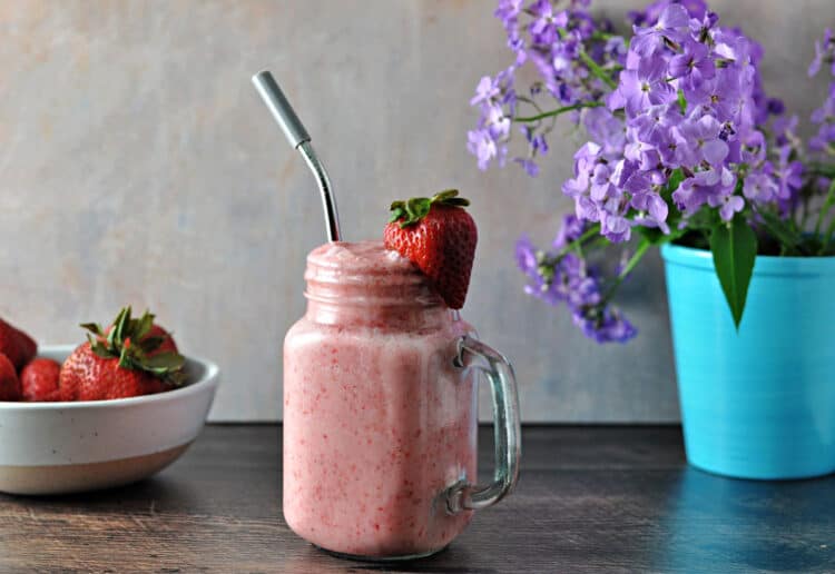 Keto strawberry smoothie with a straw, garnished with a fresh strawberries. Purple flowers and a bowl of fresh berries behind the smoothie.