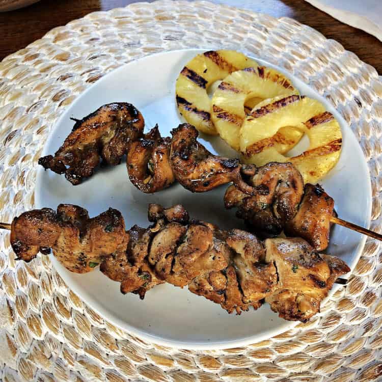 2 chicken skewers on a white plate with some grilled pineapple slices