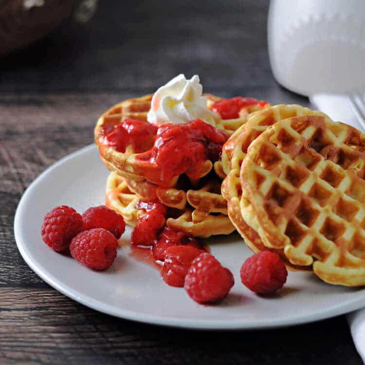 stack of 4 keto waffles (breakfast chaffles)with a little berry sauce drizzled on top and a dollop of whipped cream. 2 palin waffles lean on teh stack and some raspberries are also scattered on the plate