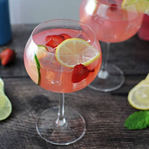 Pink gin and lemonade in a wine glass garnished with slices of lemon, lime and strawberries