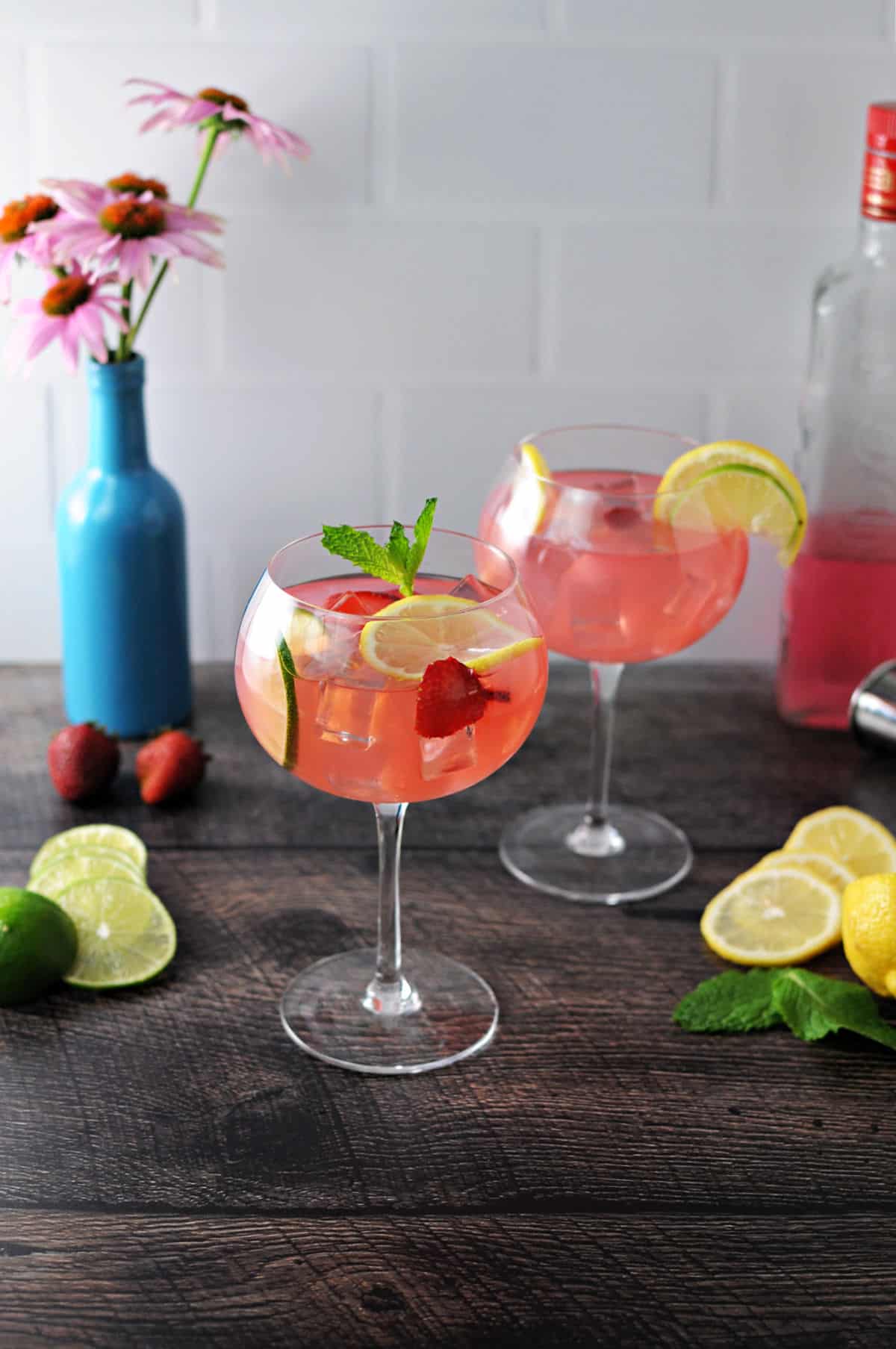 2 wine glasses of pink gin and lemonade cocktails surrounded by lemon and lime slices and a blue vase with flowers behind