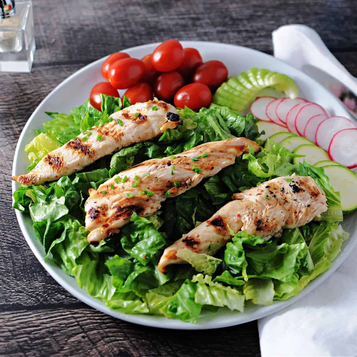 3 grilled chicken tenders on a bed of greens surrounded by salad ingredients