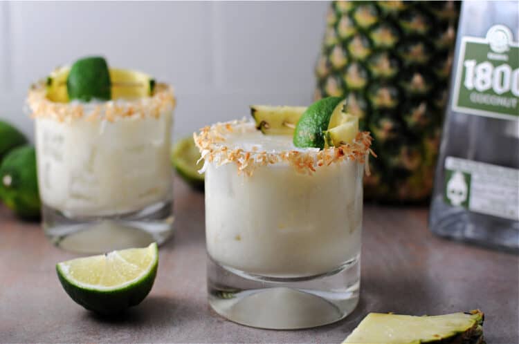 2 coconut pineapple margaritas garnished with a rim of toasted coconut. lime wedges and a whole fresh pineapple surround the area.