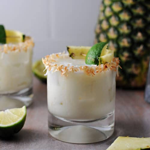 coconut pineapple margarita in a rocks glass garnished with a rim of toasted coconut and a spear of pineapple and lime