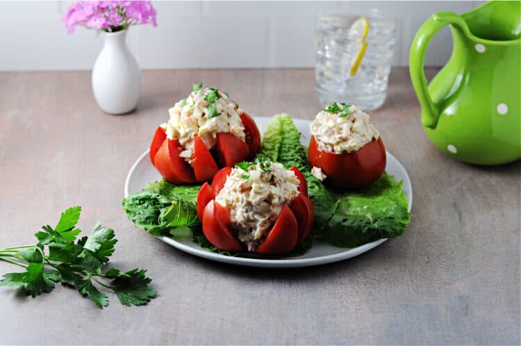 plate of 3 tuna stuffed tomatoes with a glass of water, a green pitcher and a small vase of flowers behind