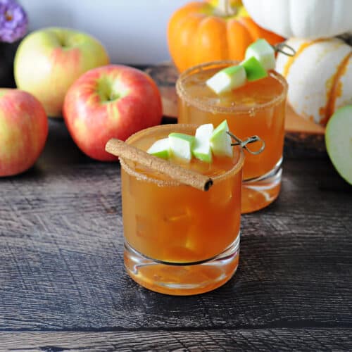 2 caramel apple cider vodka drinks garnished with apple cubes and cinnamon, whole apples behind