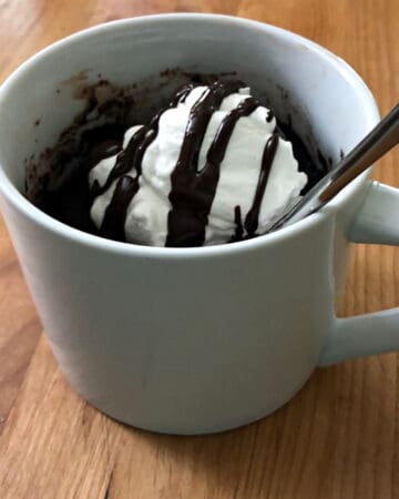 keto chocolate mug cake with whipped cream and chocolate drizzle, all in a white mug and a spoon