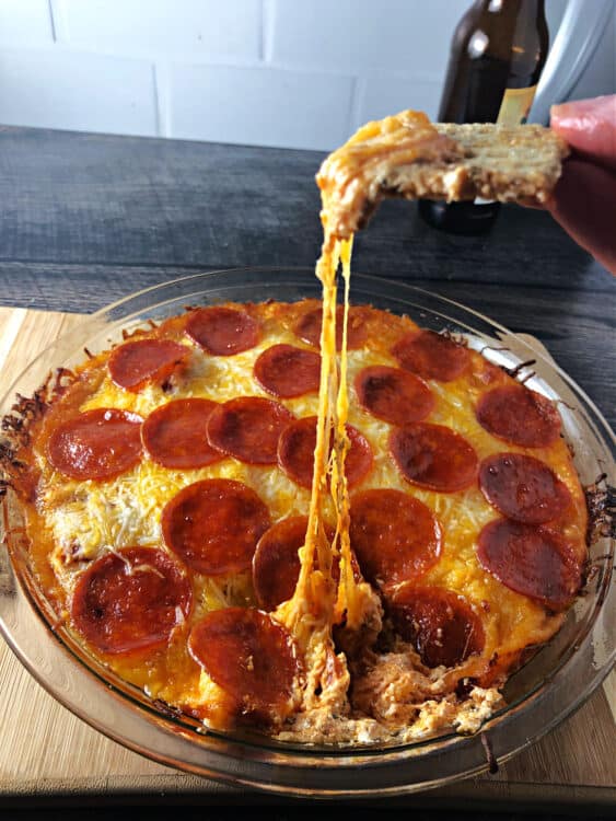 pepperoni dip showing a long pull of melted cheese between the dish and the cracker