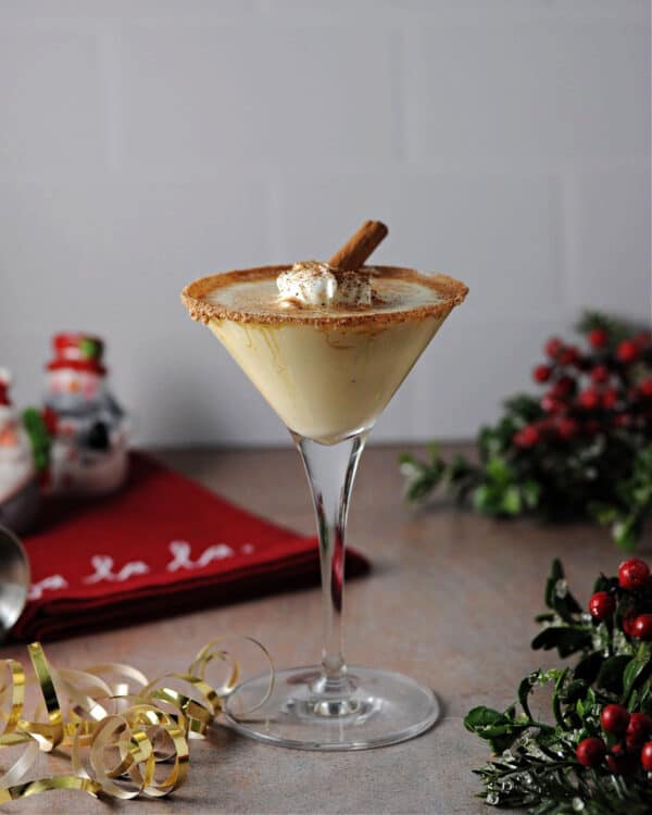 eggnog and baileys cocktail garnished with nutmeg rim, whipped cream and cinnamon stick, surrounded by Christmas holiday decor
