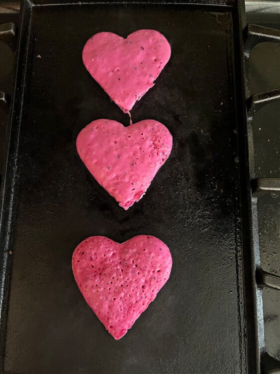 3 heart shape dragon fruit pancakes cooking on a griddle