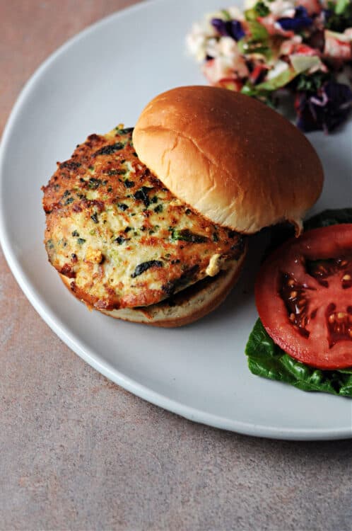 top doen view of chicken feta spinach burger on a bun, served on a plate with tomato and lettuce plus a salad on the side.