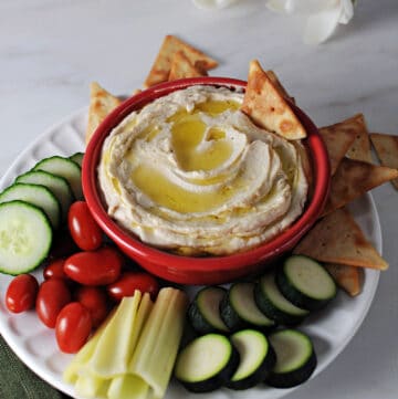 butter bean hummus in a red bowl surrounded by crackers and veggies.