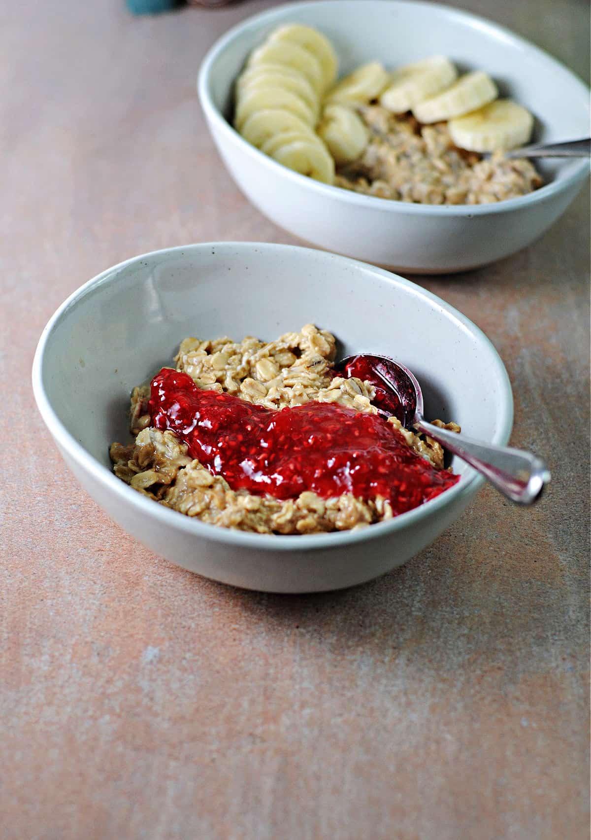 2 bowls of peanut butter overnight oats, one with bananas and the other with raspberry jam.