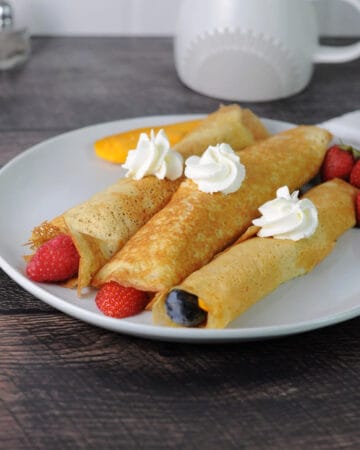 3 crepes made from pancake mix on a plate rolled with fruit and dolloped with whipped cream.