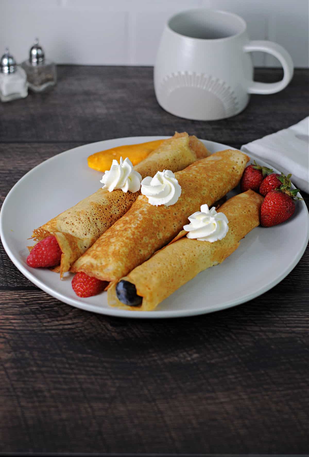 3 crepes on a plate with fruit and a dollop of whipped cream on each one.