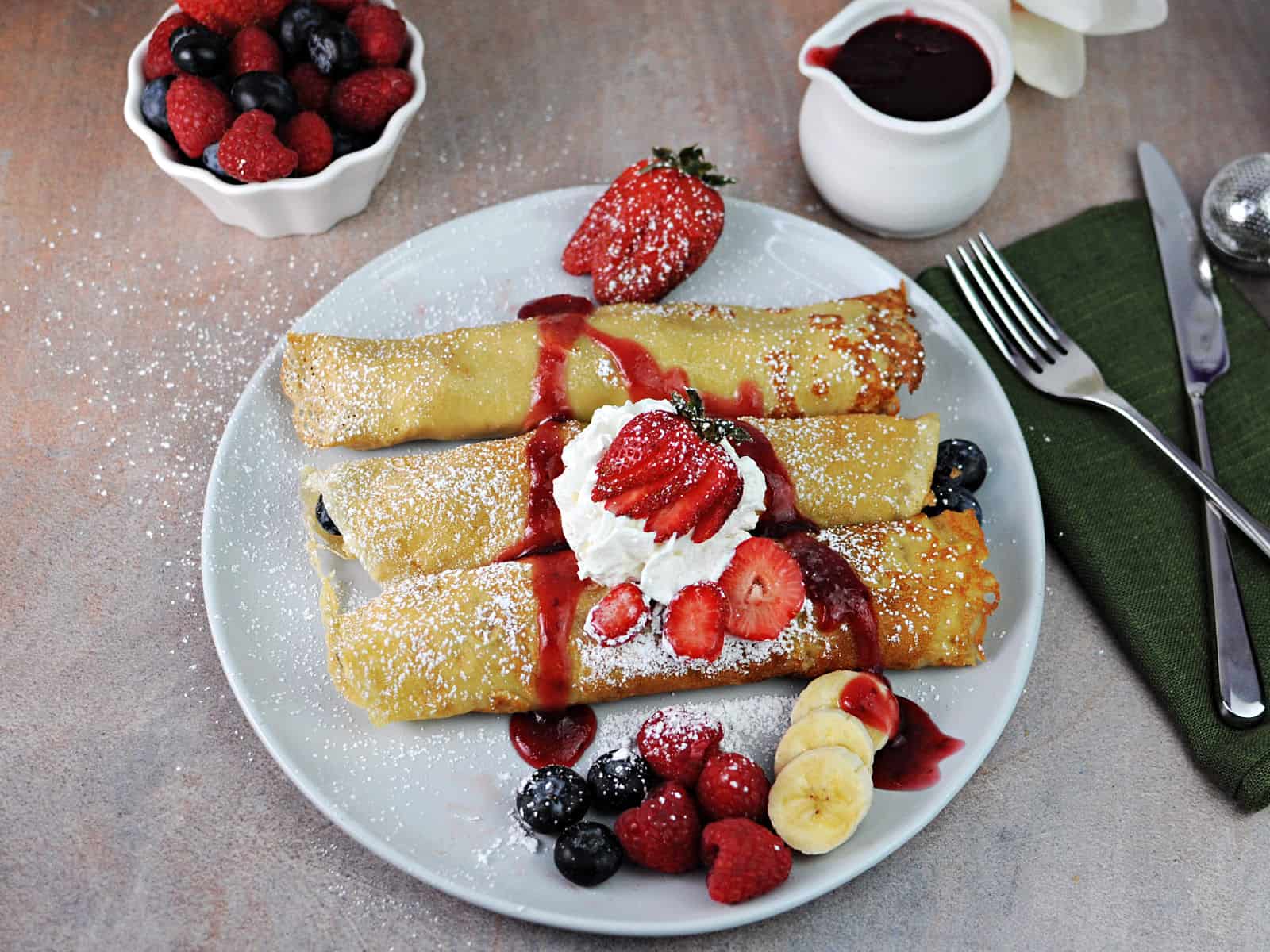 3 oat milk crepes on a plate with fruit, dusted with powdered sugar and drizzled with sauce.