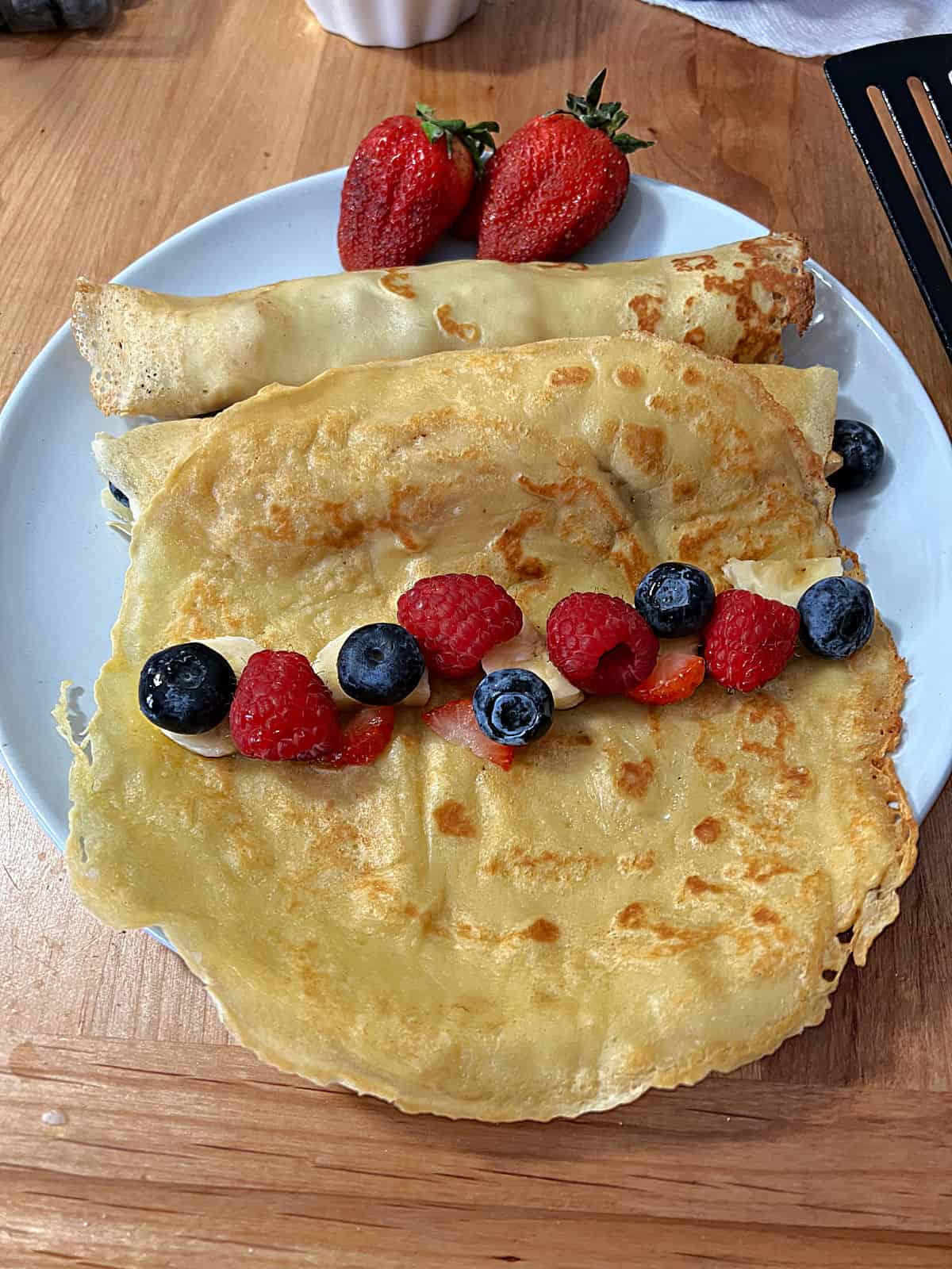 cooked crepe on a plate with berries added in a line down the middle, ready to roll up.