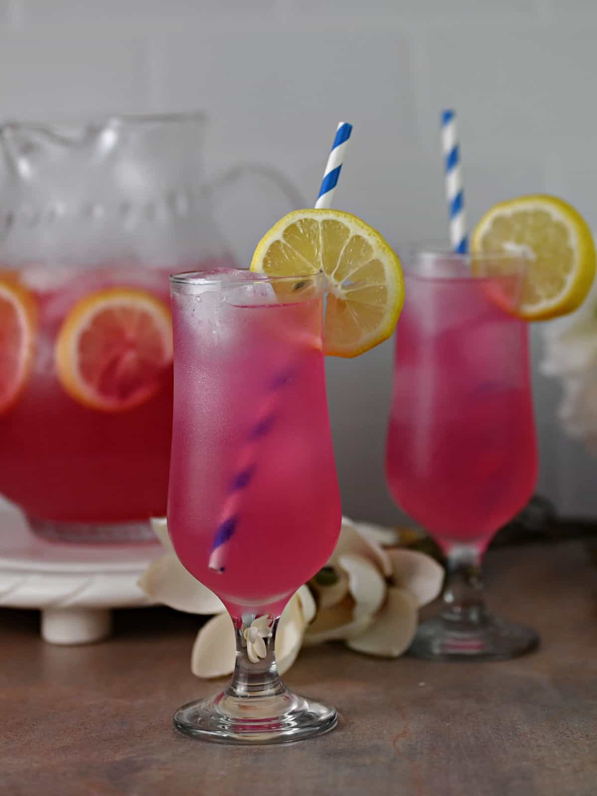 2 glasses of pink lemonade garnished with lemon and with blue and white straws, ready to drink.