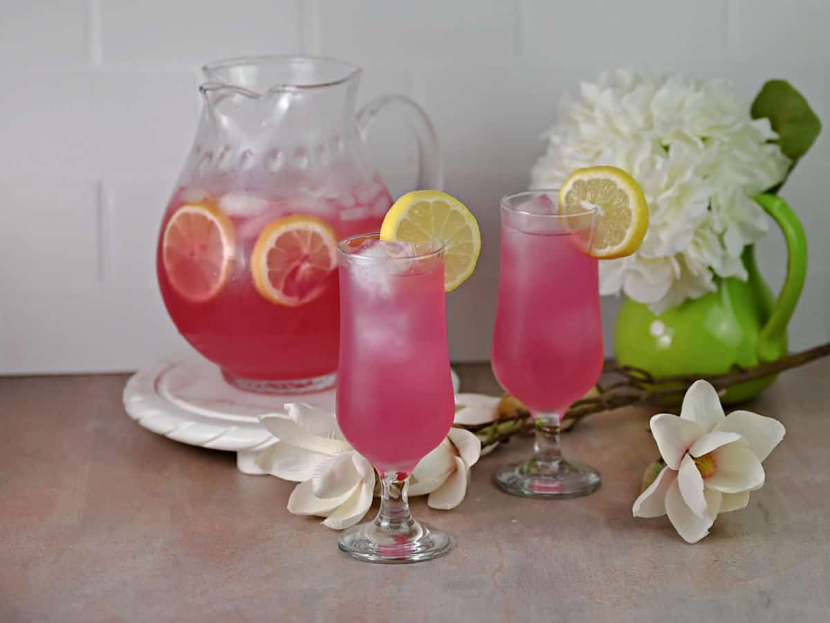2 glasses of prickly pear lemonade garnished with lemon slices, with the pitcher of lemonade behind.