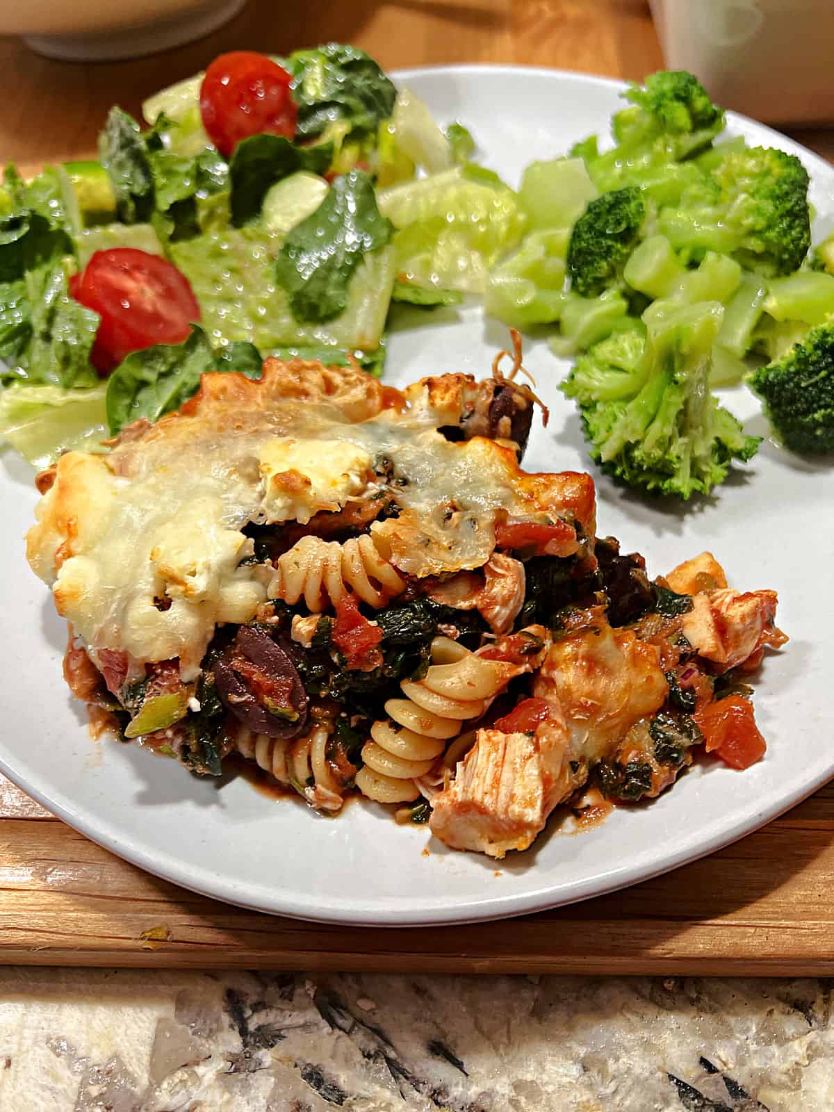 A serving of Greek chicken casserole on a plate with salad and broccoli.