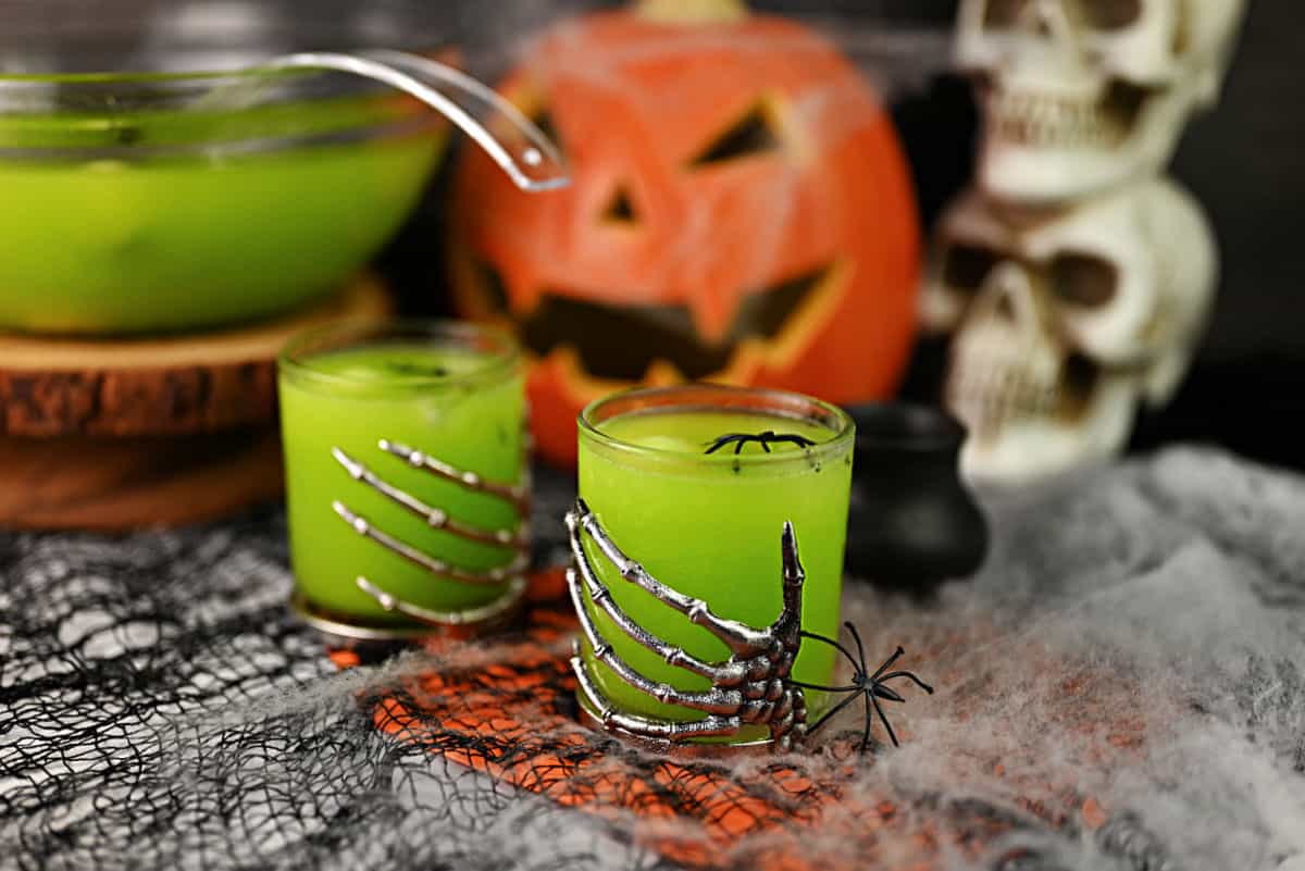 Witches brew cocktail in 2 skeleton hand glasses with a punch bowl filled with more behind.