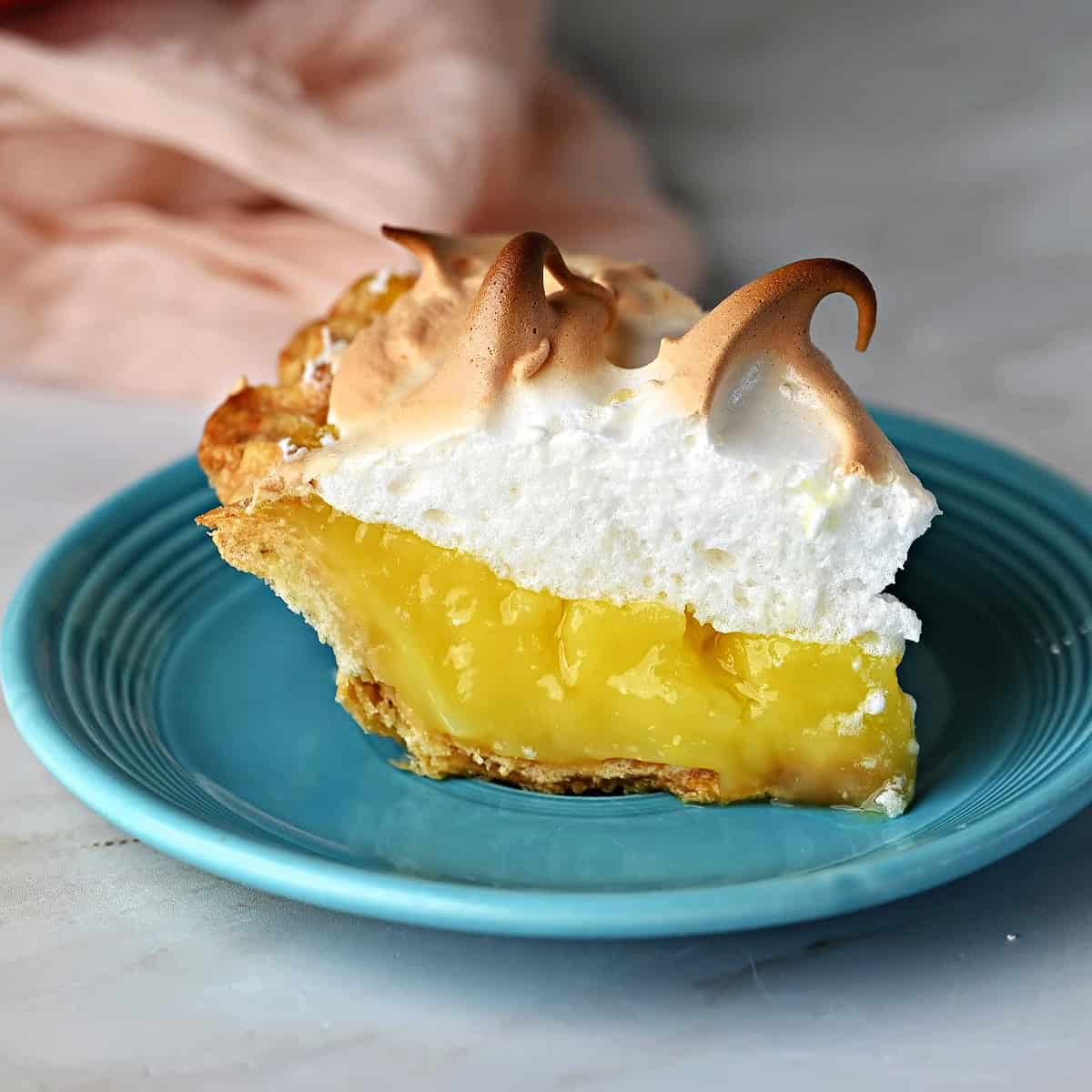A slice of lemon meringue pie, on a small blue plate, ready to eat.